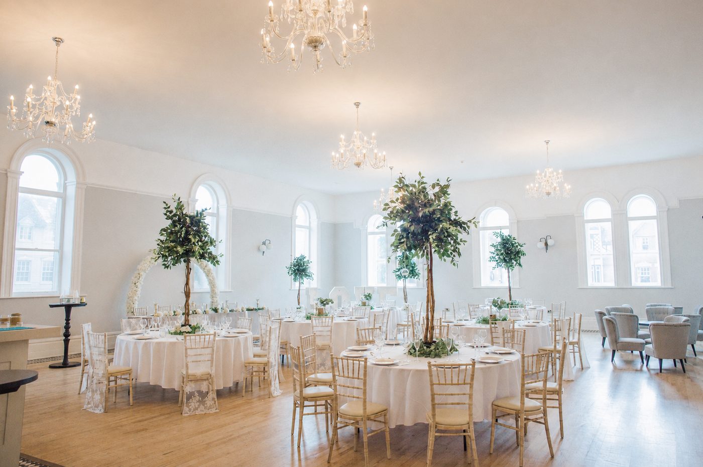 The Ballroom dressed for a wedding breakfast, with round tables, tree centrepieces and chandeliers beneath tall arched windows